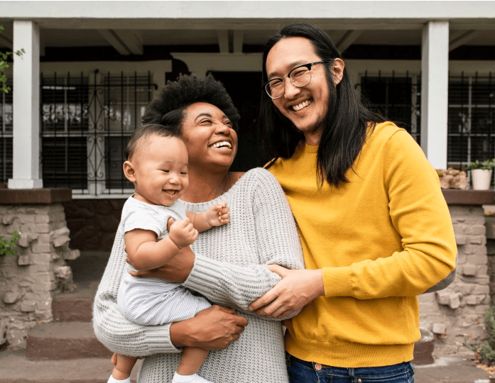 image d'un jeune couple riant devant leur maison tout en tenant leur jeune enfant dans ses bras